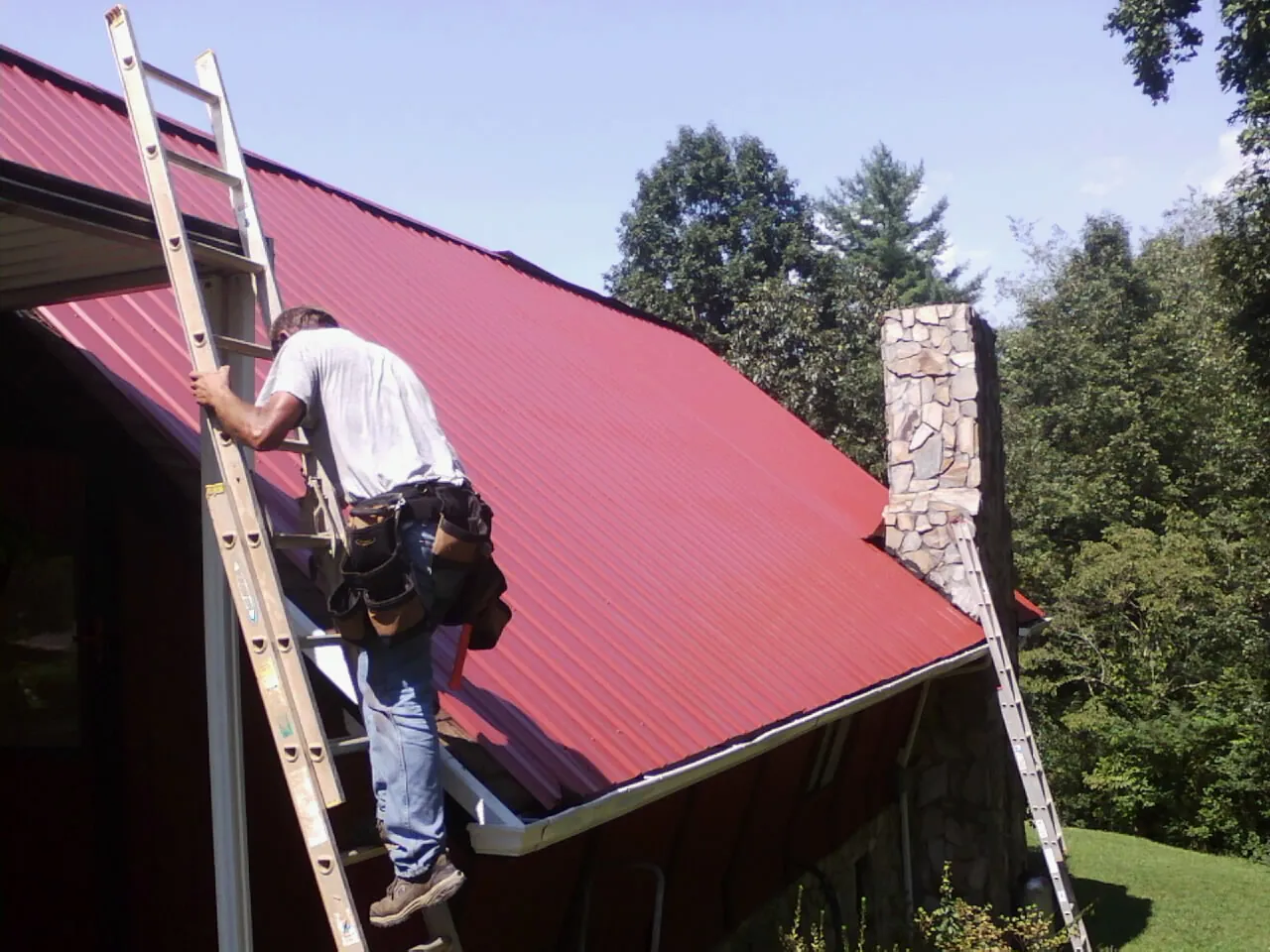 Roofer on a ladder beside a red metal roof and stone chimney — Soffit & Fascia Repair by Roofing Brothers in Ellwood City